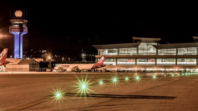Pátio do Aeroporto Internacional de São Paulo, em Guarulhos, à noite, com a torre de controle iluminada, o terminal de passageiros ao fundo e luzes verdes de balizamento na pista. Imagem de arquivo.