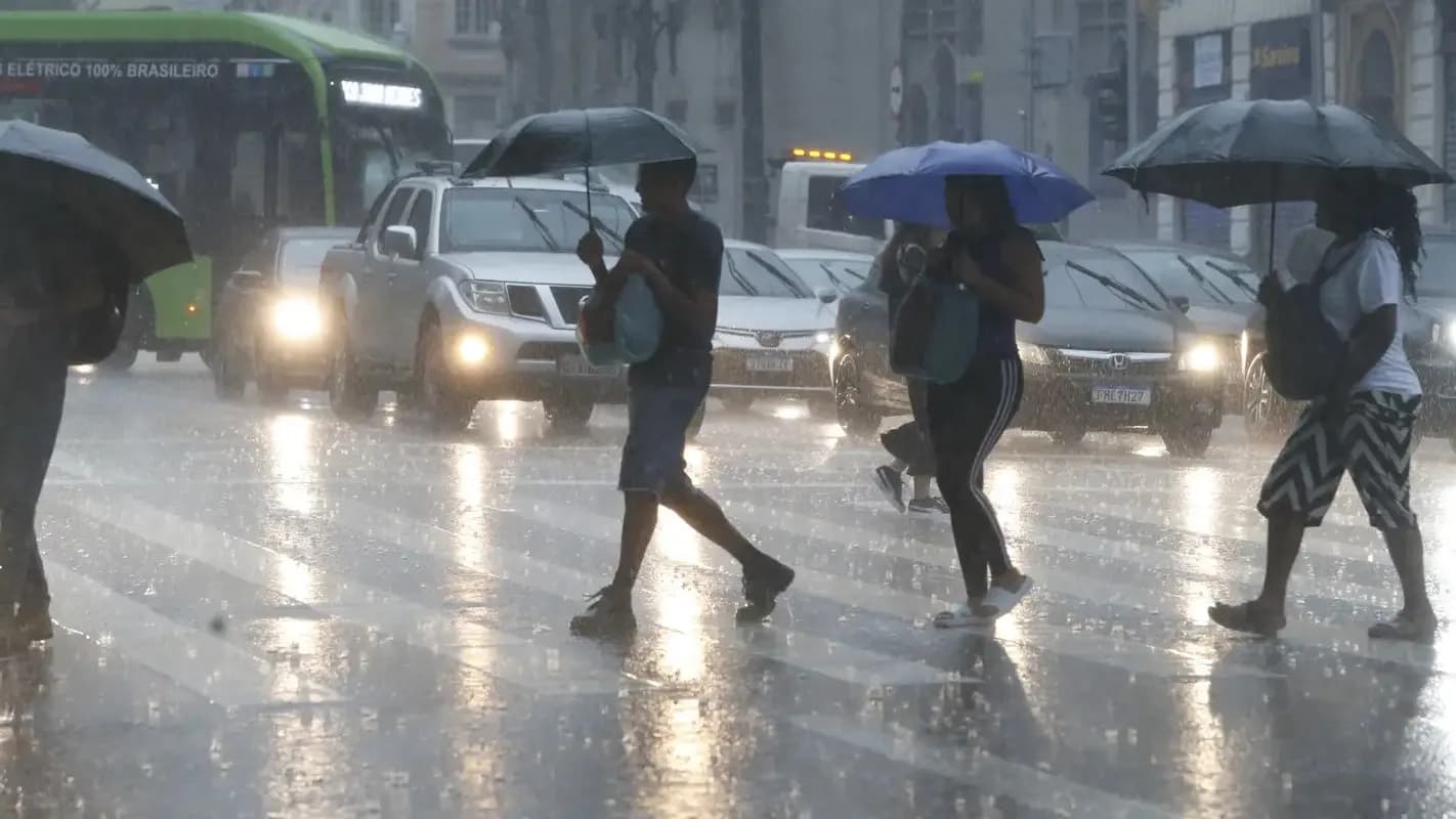 Pedestres caminham sob chuva forte com guarda-chuvas em via de São Paulo