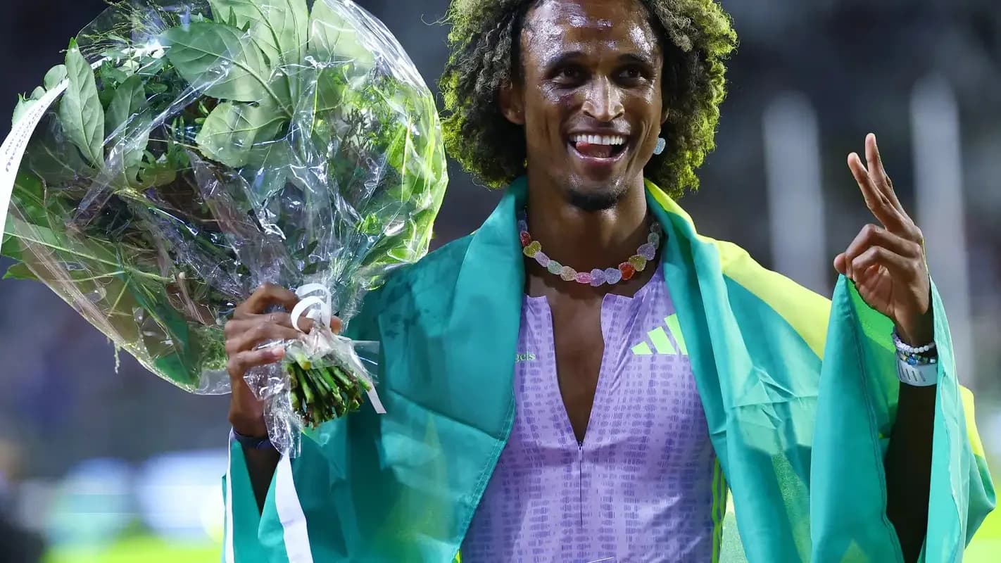 Alison dos Santos sorri e comemora com buquê de flores e bandeira do Brasil nos ombros