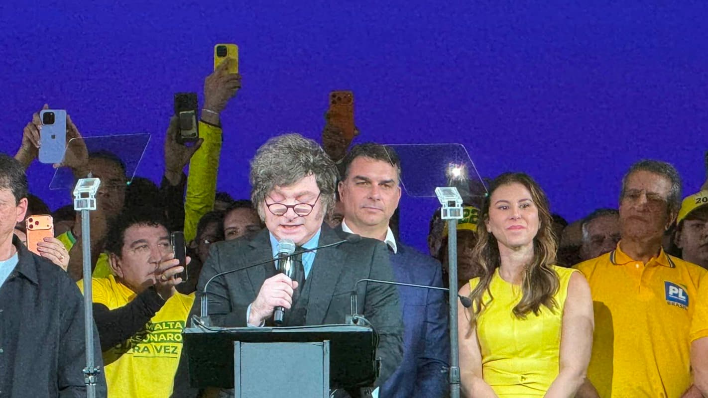 Javier Milei discursa ao microfone durante convenção do PL em São Paulo, com Flávio Bolsonaro ao fundo