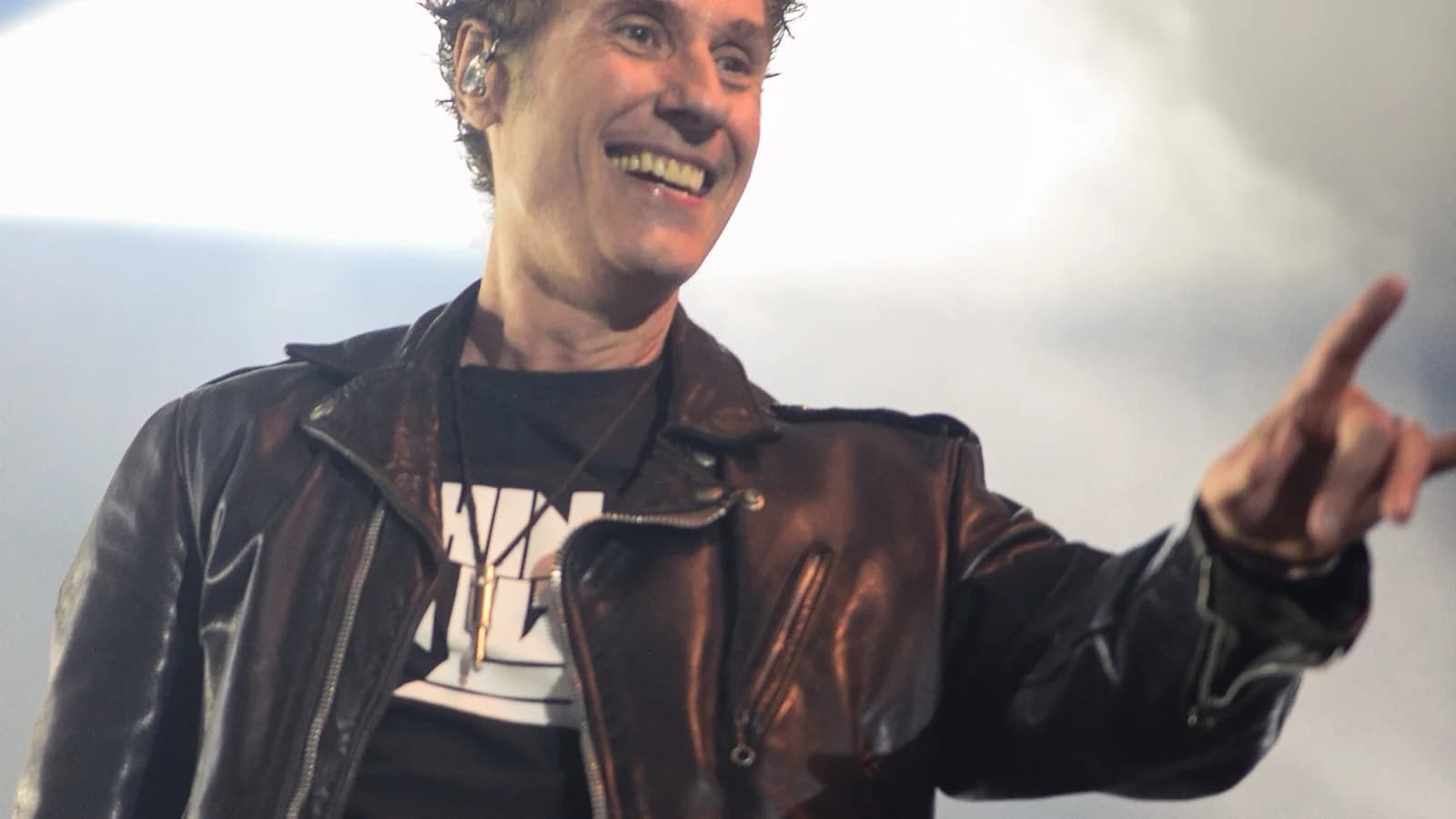 Dinho Ouro Preto sorri no palco, usando jaqueta de couro preta, apontando para o público durante show