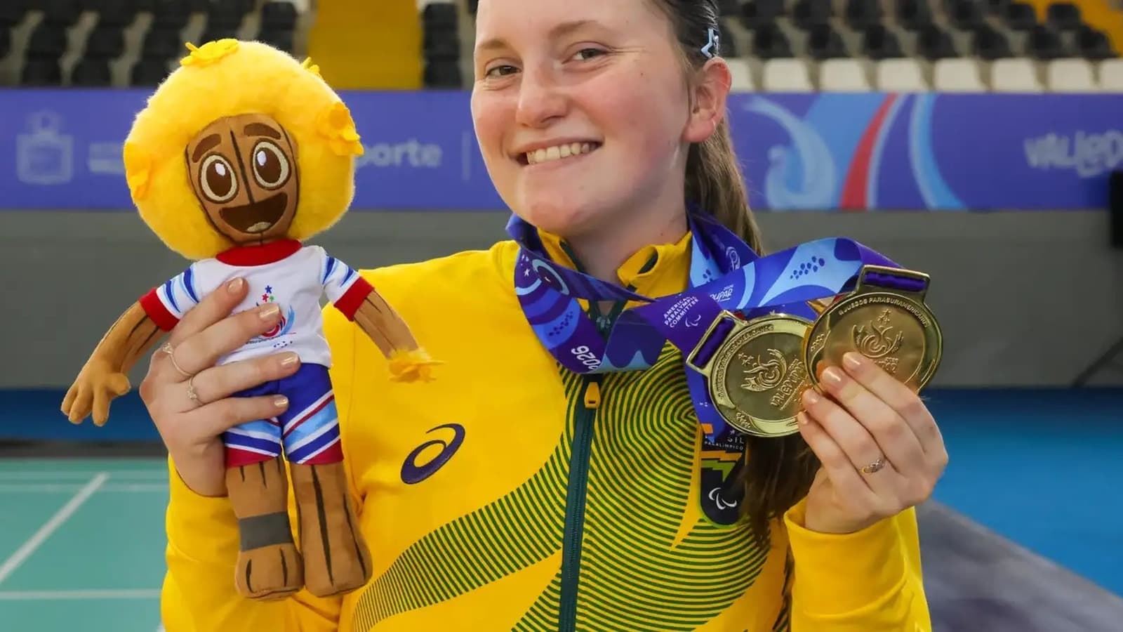 A jogadora de parabadminton Kauana Beckenkamp, de agasalho amarelo da delegação brasileira, sorri segurando duas medalhas de ouro e um boneco de mascote — imagem de arquivo de competição anterior