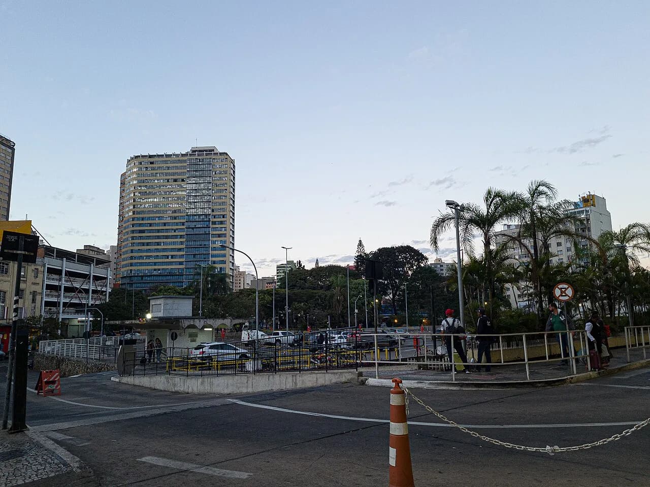 Vista externa da área de embarque e desembarque da Rodoviária de Belo Horizonte ao entardecer, com passageiros aguardando junto às grades de acesso, cone de sinalização em primeiro plano e prédios do centro da cidade ao fundo