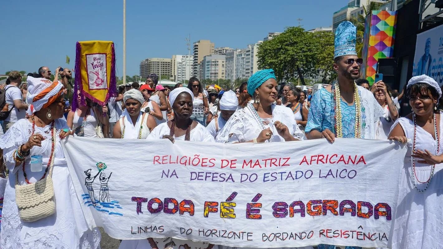 Grupo de pessoas de religiões de matriz africana em marcha, segurando faixa com os dizeres 'Toda fé é sagrada'