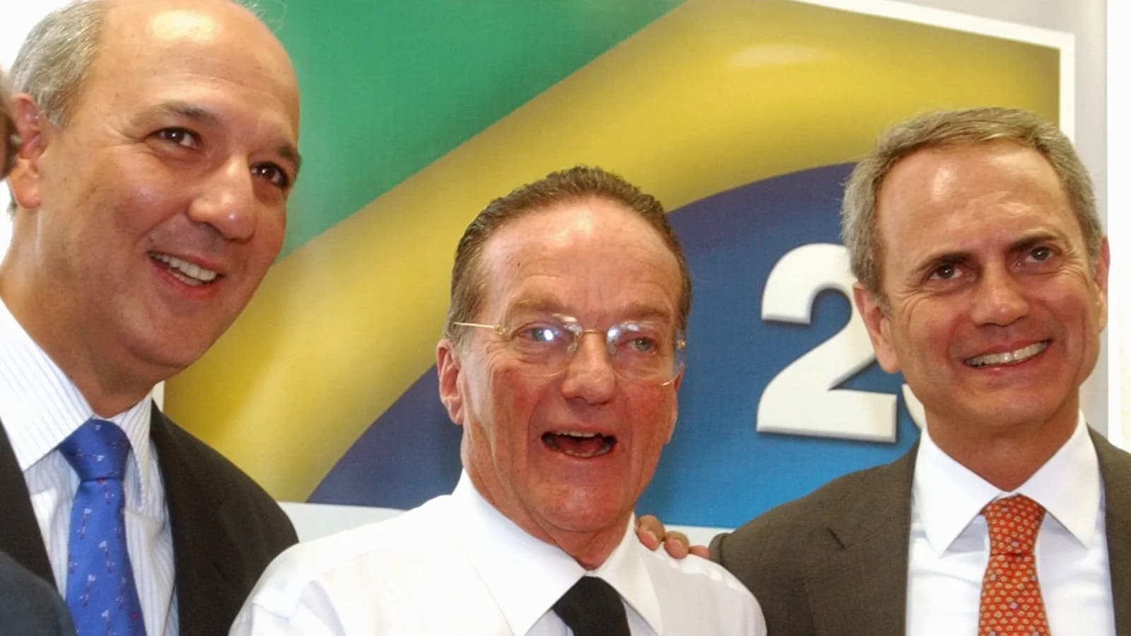 José Roberto Arruda sorrindo ao lado de outros dois políticos, com bandeira do Brasil ao fundo