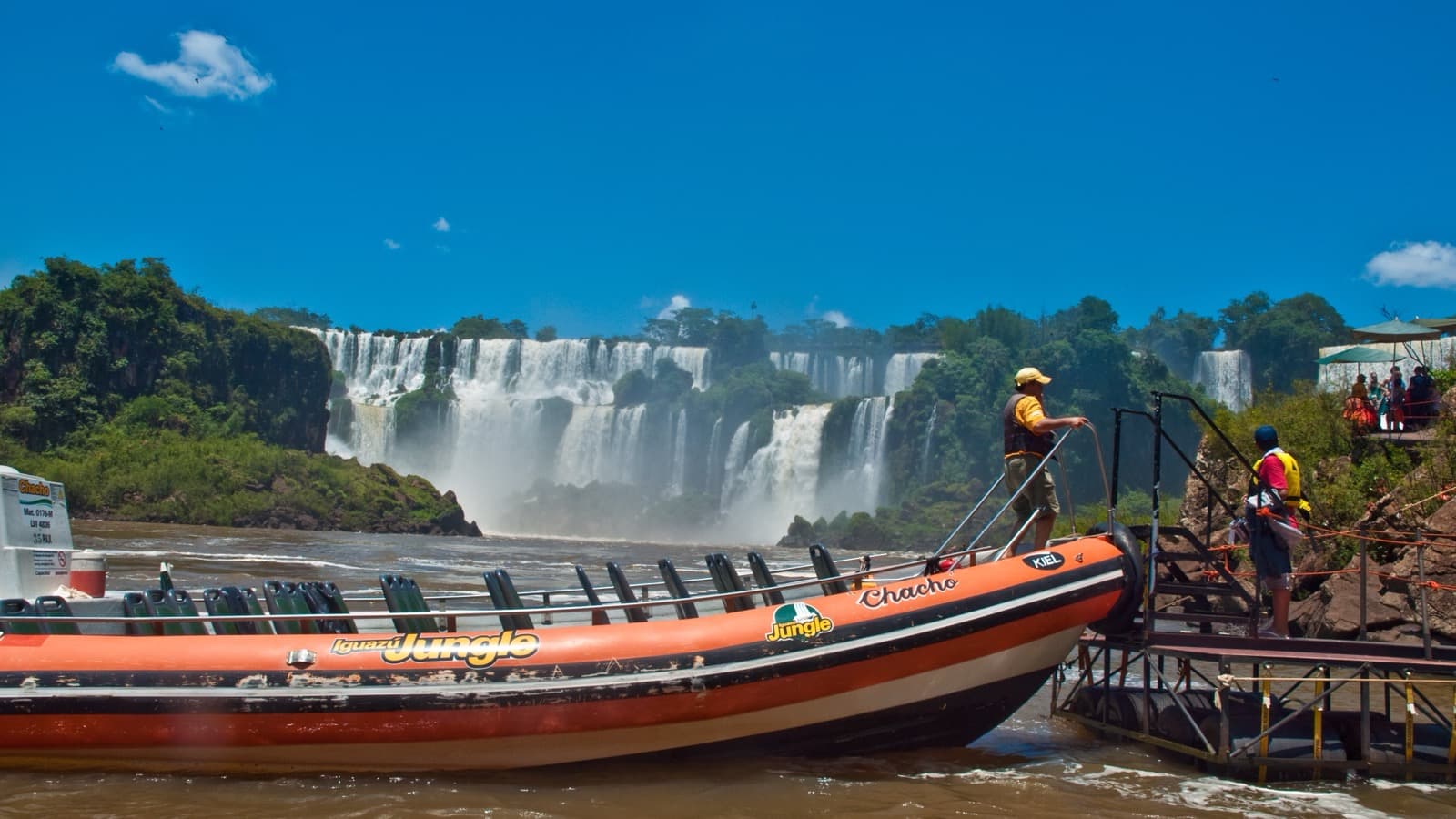 Barco de passeio turístico com tripulantes de colete salva-vidas nas Cataratas do Iguaçu, com as quedas d'água ao fundo