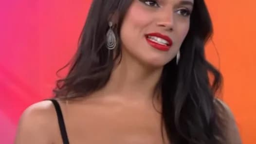 Alane Dias sorri em entrevista de televisão, com fundo colorido em tons de rosa e laranja