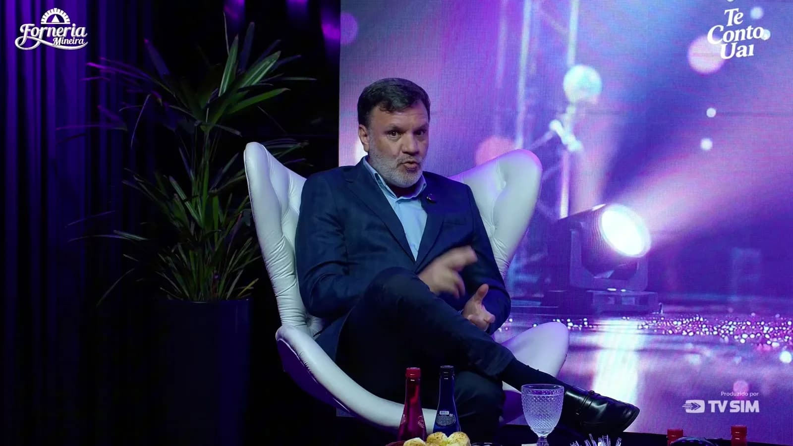 Alessandro Feijó fala olhando para o lado durante entrevista ao programa Te Conto, Uai, em estúdio com luzes roxas