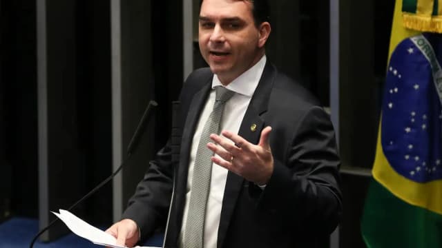 Flávio Bolsonaro discursa em um púlpito, com a bandeira do Brasil ao fundo