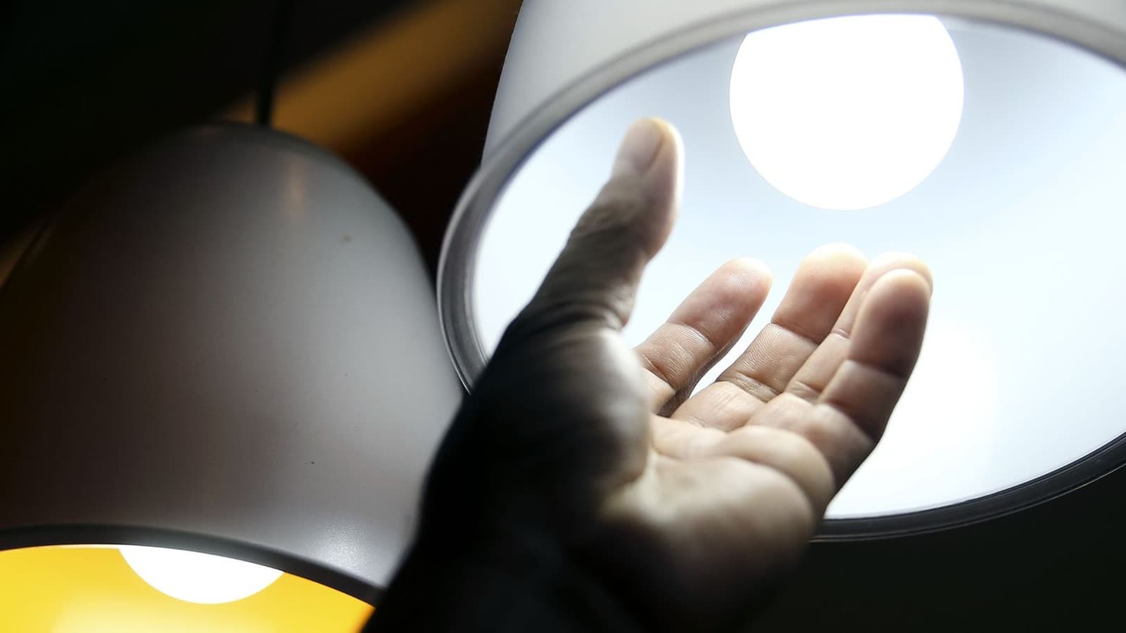 Mão de uma pessoa diante de duas luminárias acesas, uma com luz amarela e outra com luz branca; imagem de arquivo sobre consumo de energia elétrica