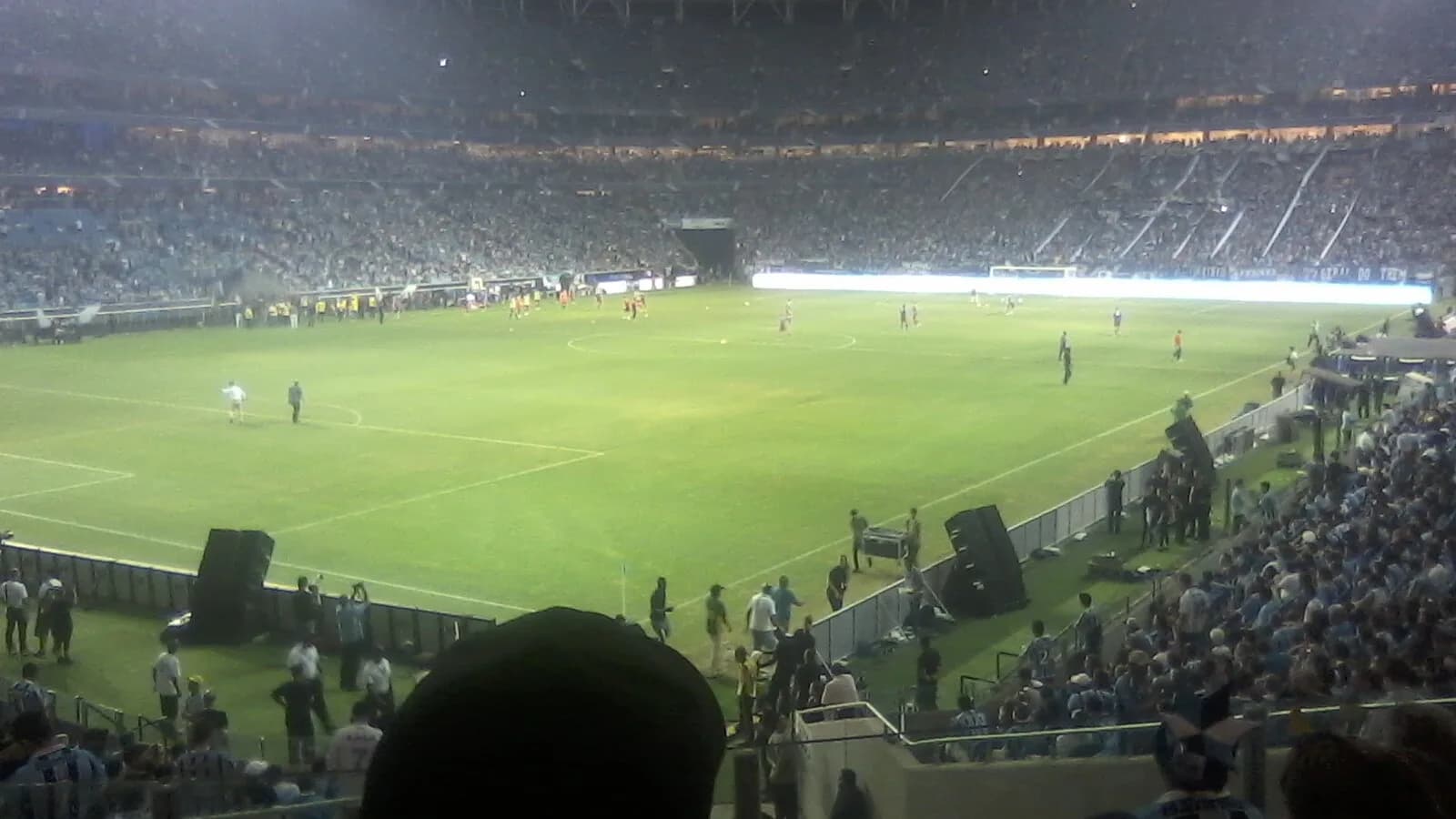 Interior da Arena do Grêmio lotada durante uma partida, com o gramado iluminado
