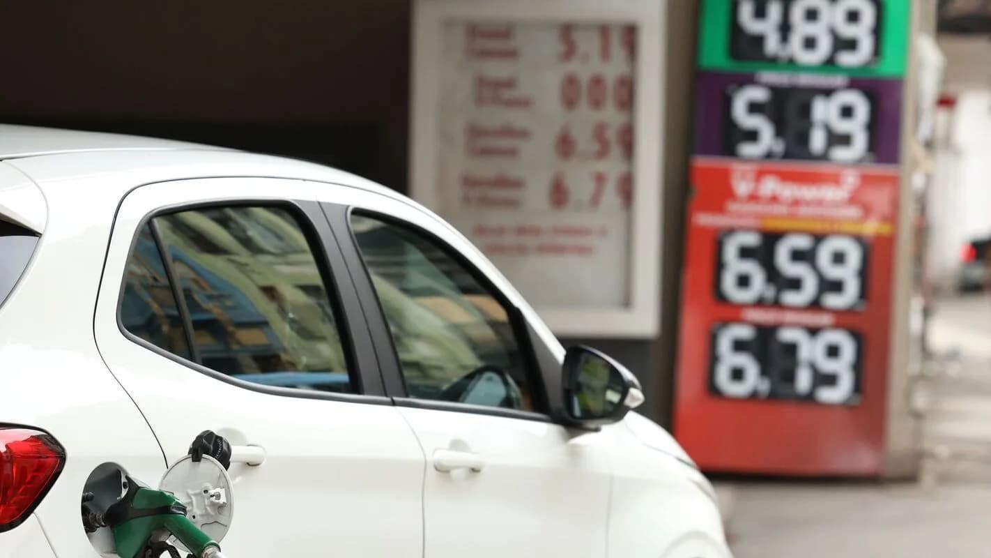 Carro branco é abastecido em posto de combustível com placas de preço da gasolina ao fundo
