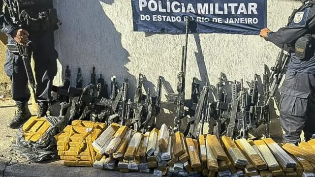 Dois policiais militares do Rio de Janeiro perfilados junto a fuzis e tabletes de drogas apreendidos, em frente a faixa da Polícia Militar do Estado do Rio de Janeiro