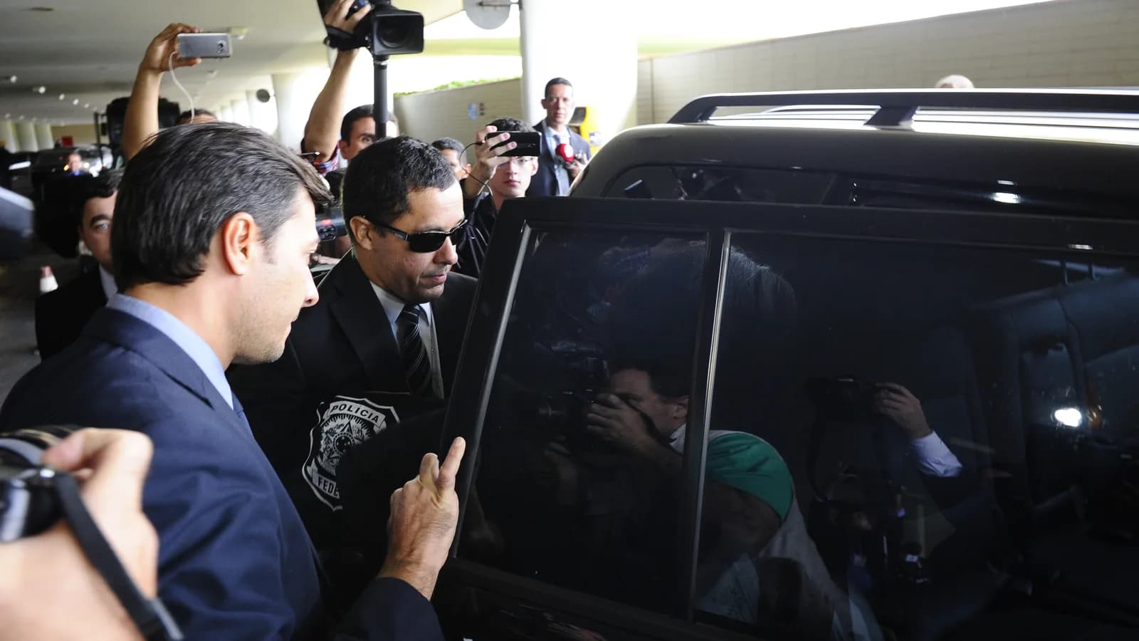 Agente da Polícia Federal em operação, cercado por jornalistas, ao lado de um veículo