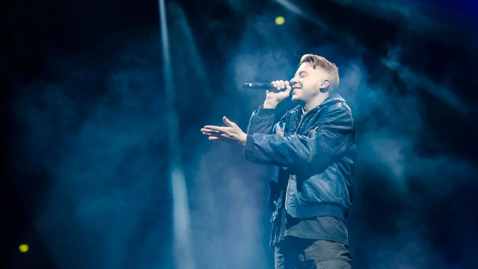 Macklemore canta ao microfone em show, com luzes azuis de palco ao fundo (foto de arquivo)