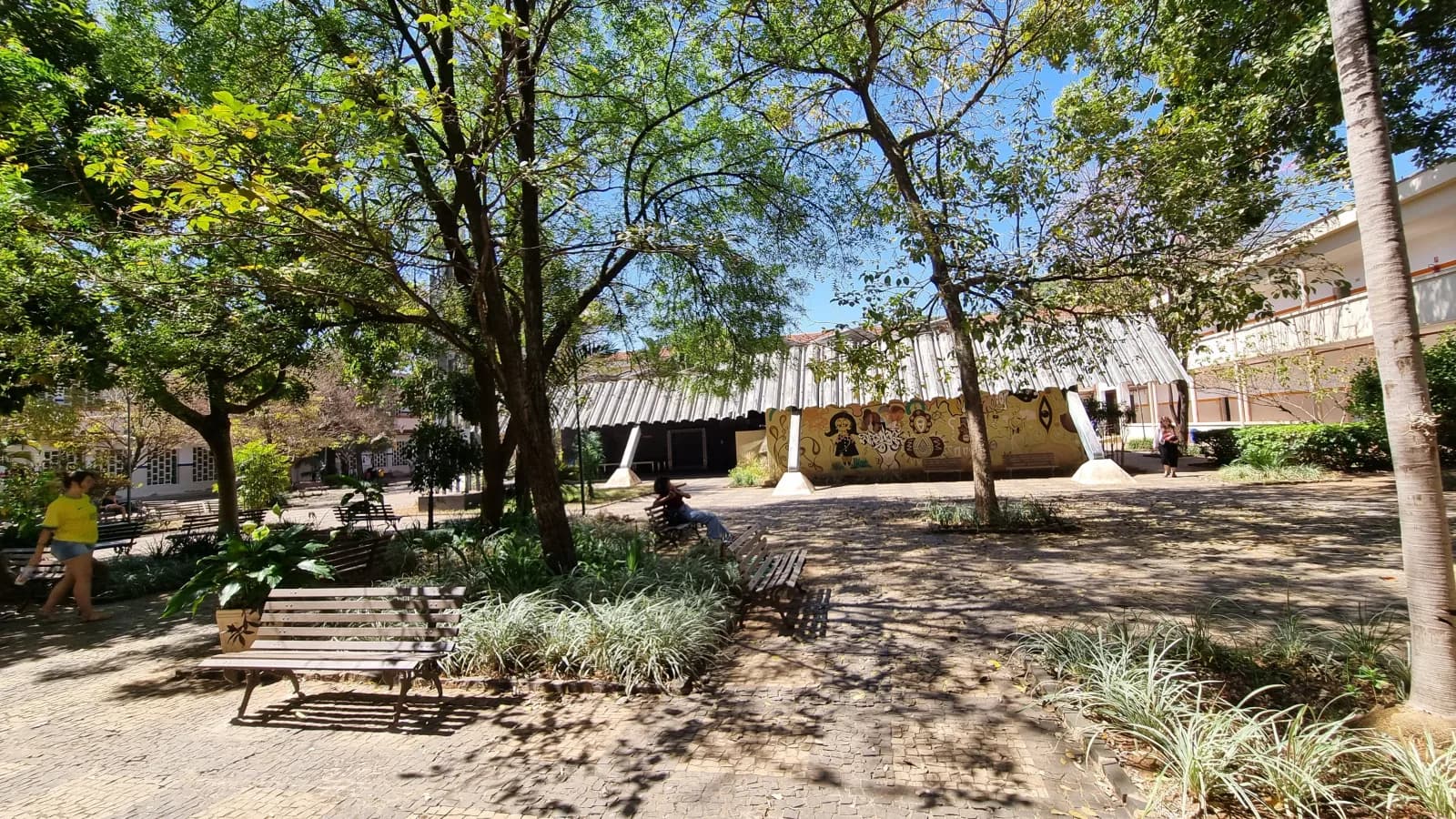 Pátio arborizado de campus de instituto federal com estudantes caminhando entre bancos, imagem ilustrativa