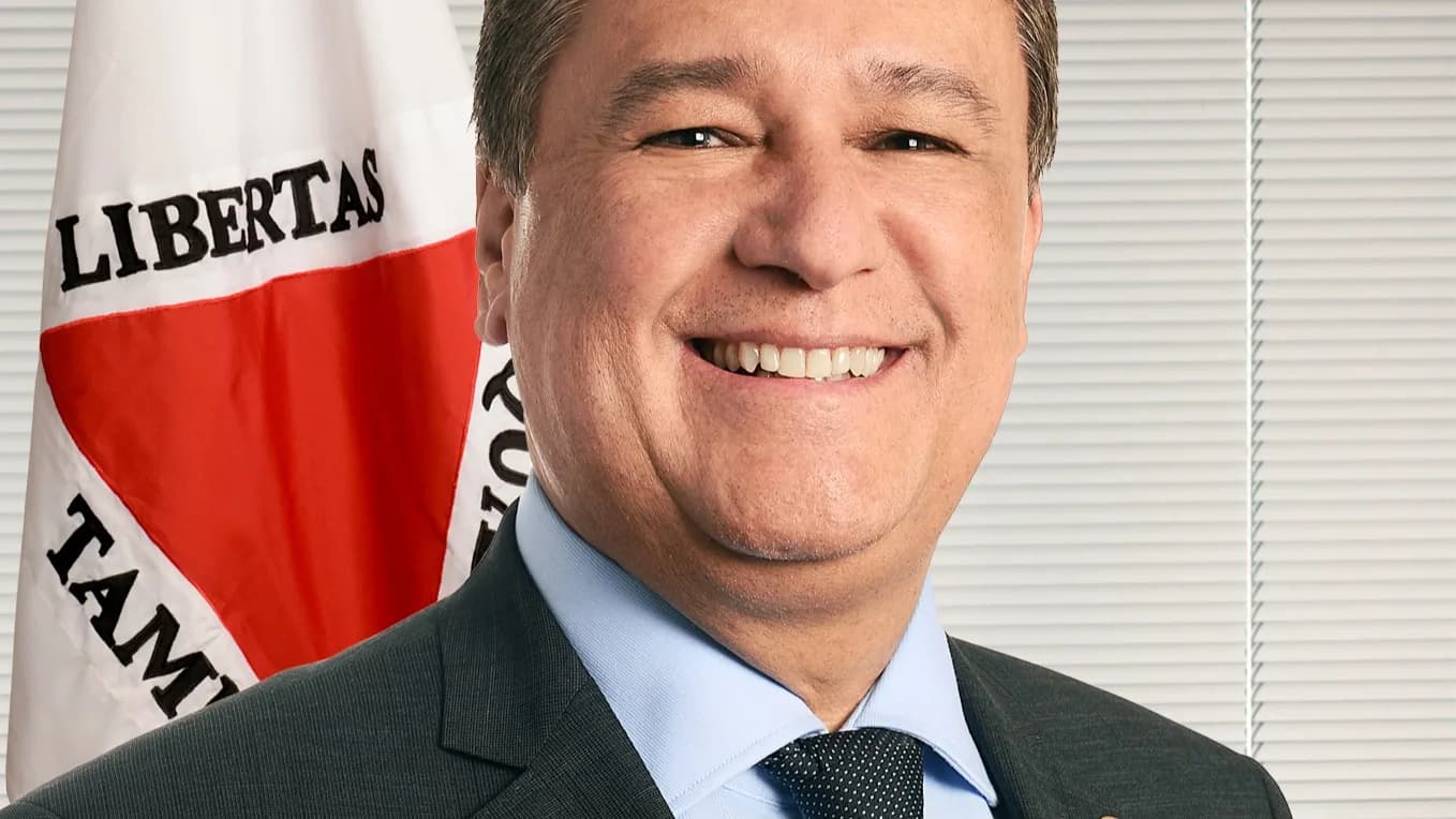 O senador Carlos Viana (PSD-MG) sorrindo, em retrato oficial, com a bandeira de Minas Gerais ao fundo