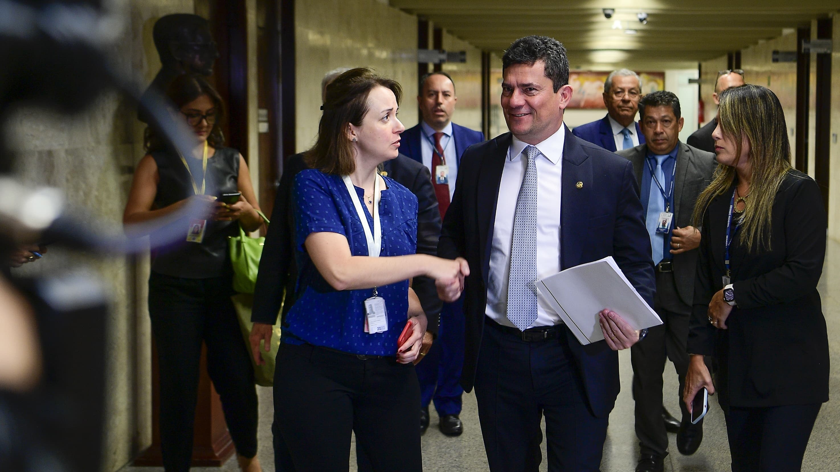 Senador Sergio Moro caminha por corredor do Senado cumprimentando assessora, cercado de outras pessoas