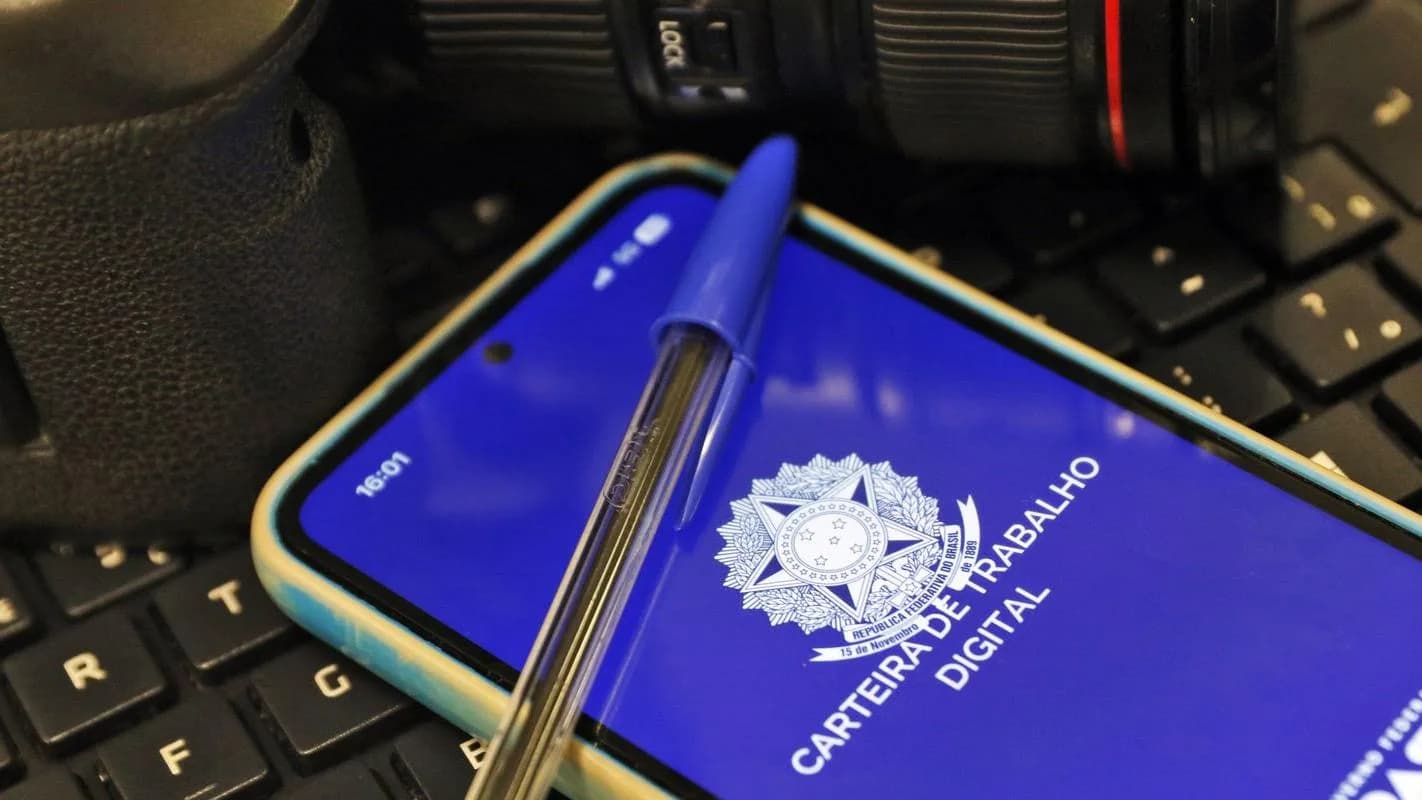 Carteira de Trabalho Digital aberta em um celular, ao lado de câmera fotográfica e teclado