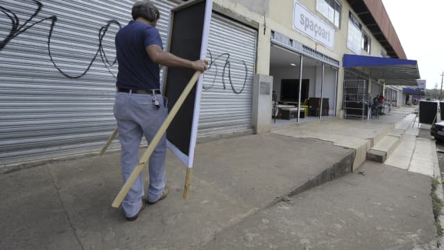 Homem instala placa de propaganda eleitoral em calçada de rua comercial