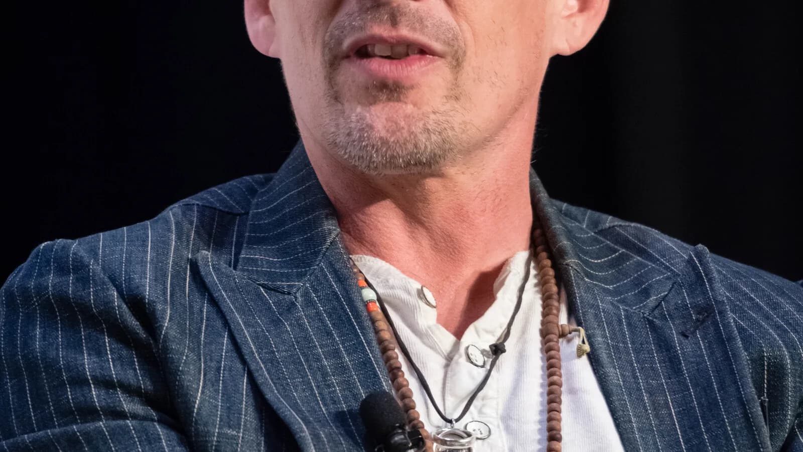 Ethan Hawke em close durante entrevista, com colar e blazer listrado