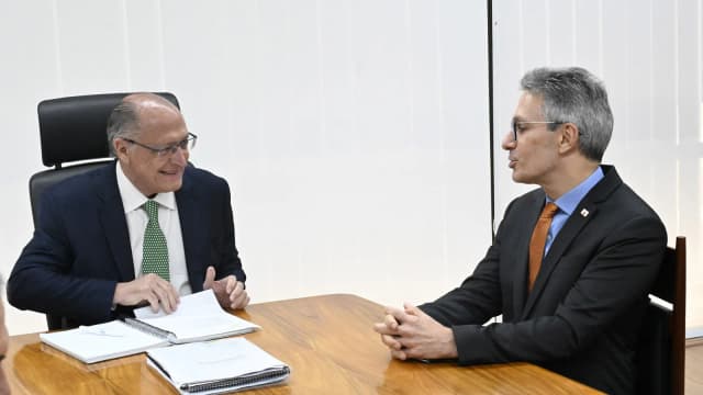 Romeu Zema em reunião de trabalho, sentado à mesa com outro homem de terno