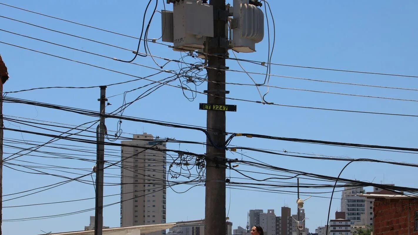 Poste de rede elétrica com transformadores e fiação em área urbana, prédios ao fundo (foto de arquivo, contexto)