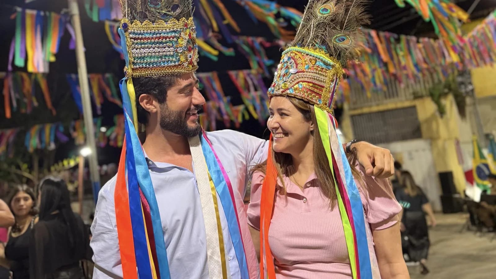 Gabriel Azevedo, candidato do MDB ao governo de Minas Gerais, sorri ao lado de uma mulher, ambos usando coroas de festa junina com penas de pavão