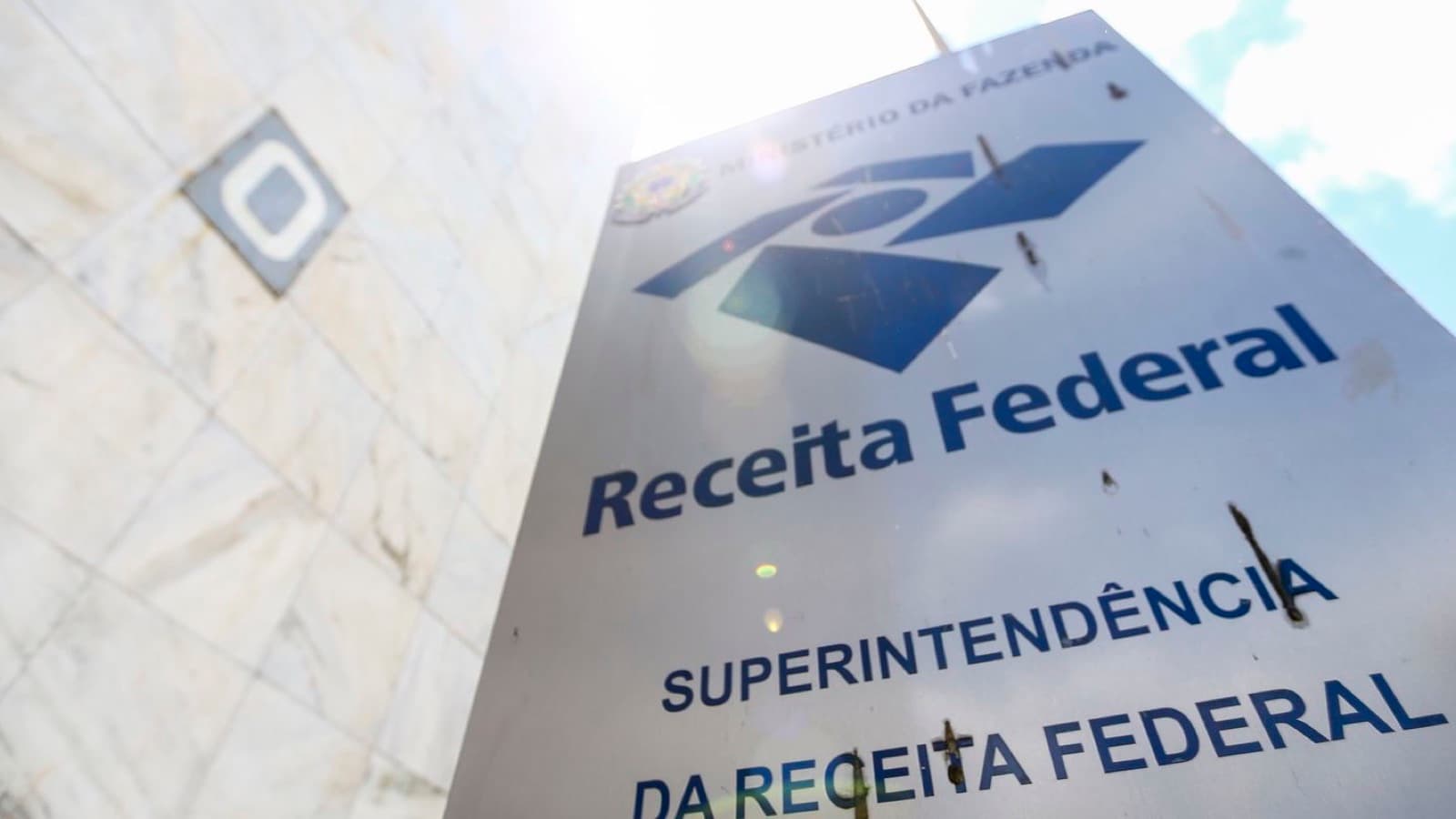 Placa da Superintendência da Receita Federal, com o logotipo do órgão e a inscrição Ministério da Fazenda, fotografada de baixo para cima contra o céu