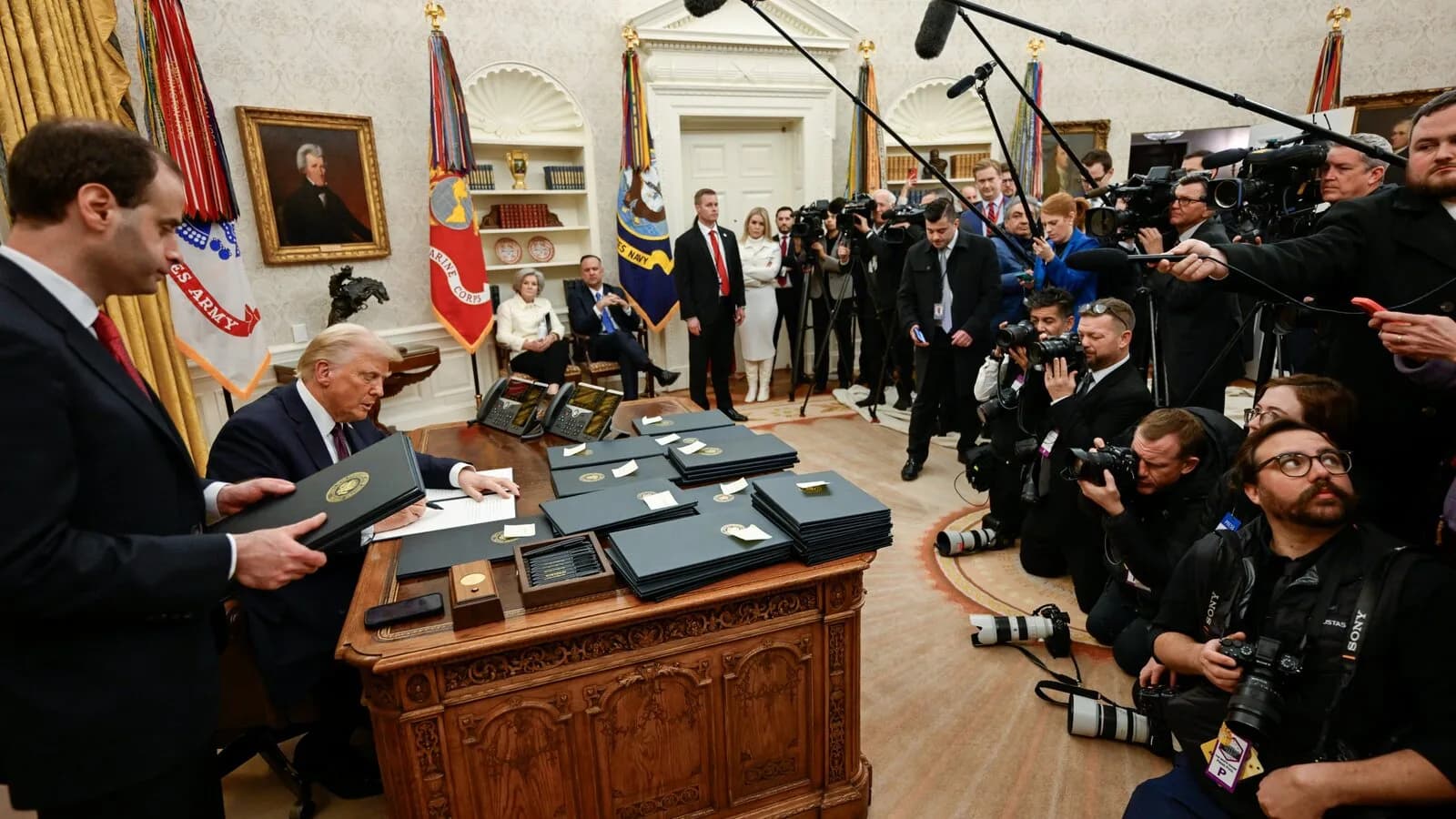 Donald Trump assina documentos em sua mesa no Salão Oval, cercado por fotógrafos e assessores