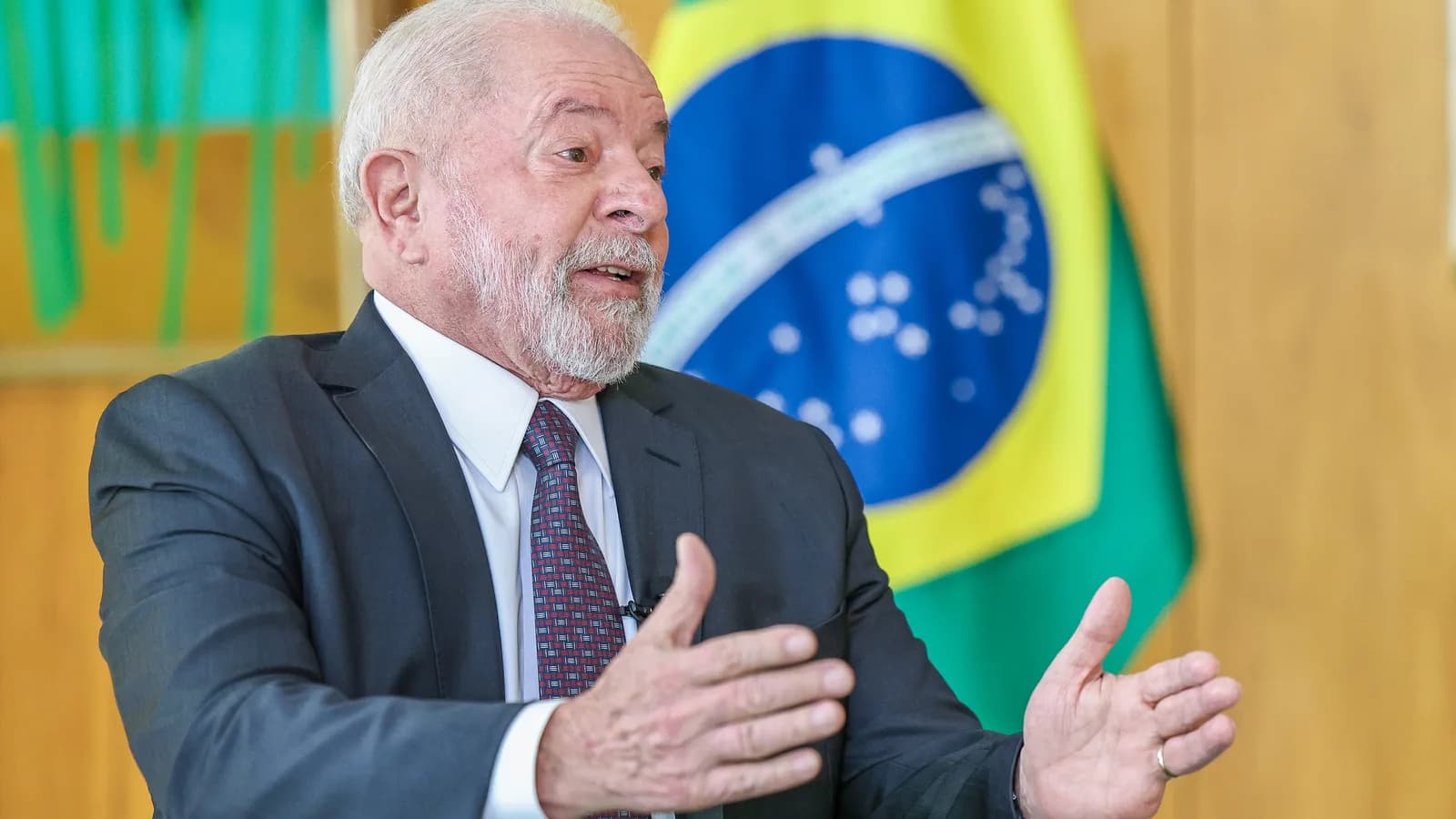 O presidente Lula gesticula durante entrevista, com a bandeira do Brasil ao fundo