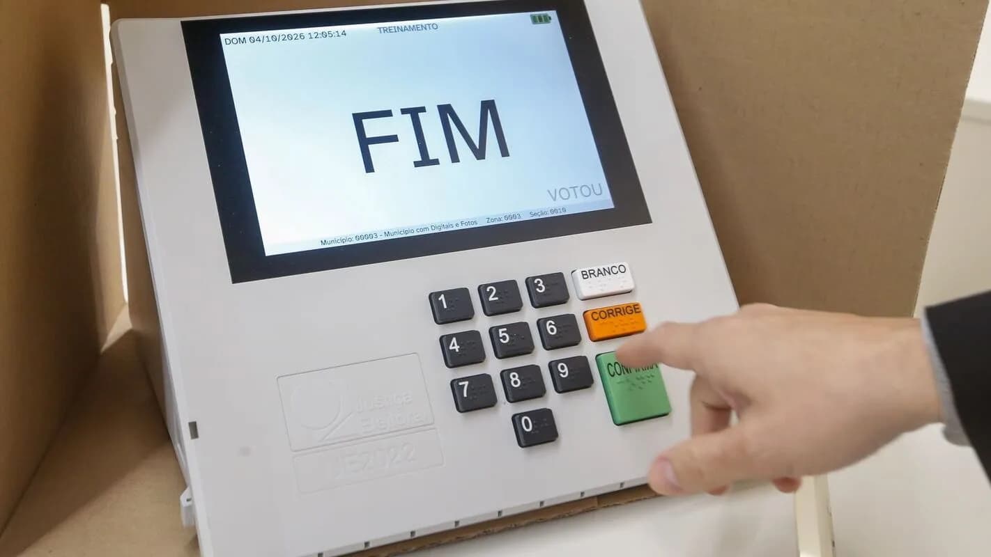 Mão de eleitor digita na urna eletrônica, com tela mostrando a palavra FIM após o voto