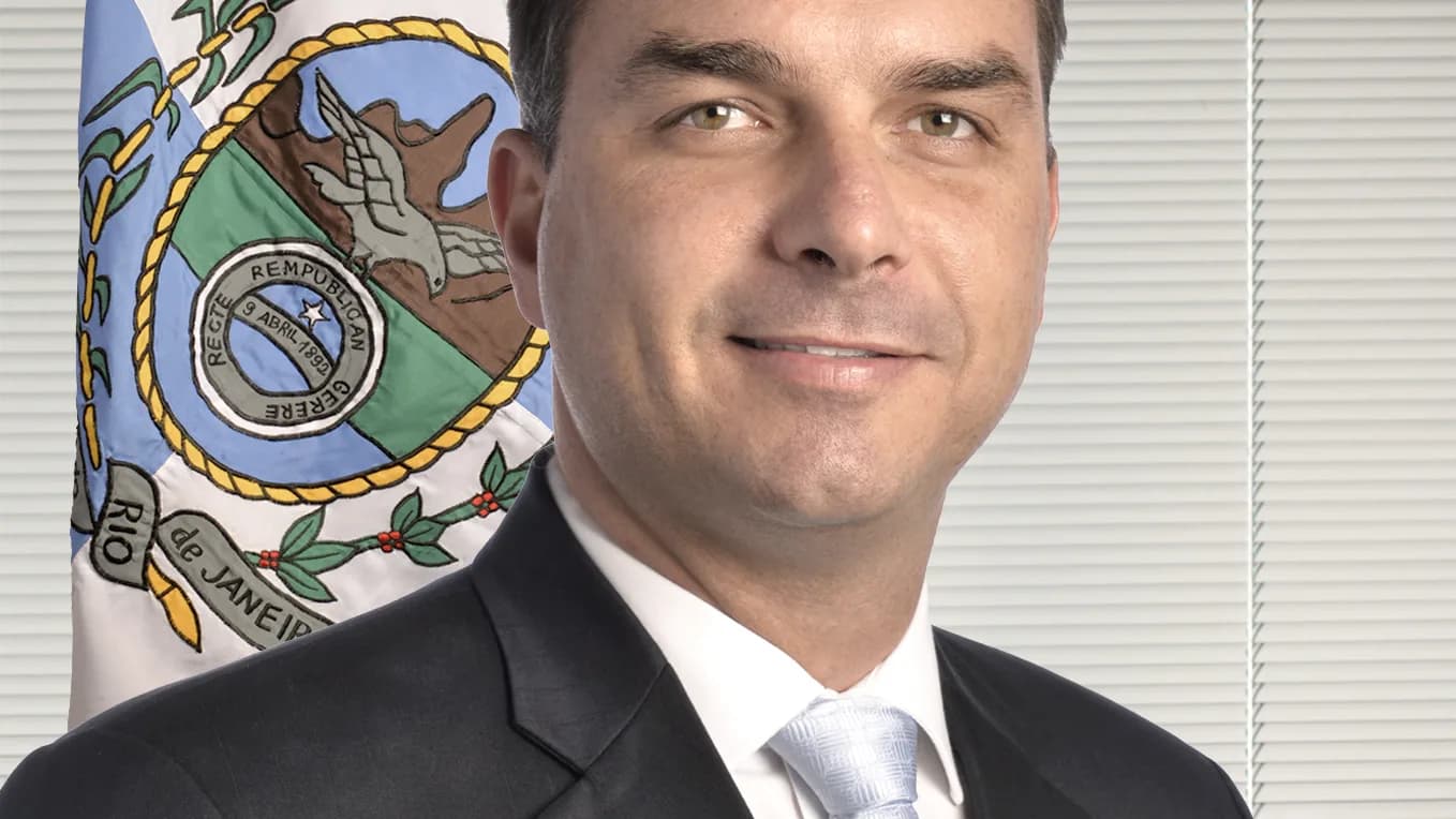Flávio Bolsonaro em retrato oficial, de terno escuro, com bandeira do Rio de Janeiro ao fundo
