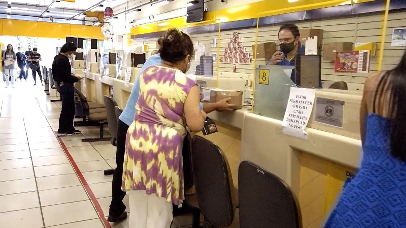 Cliente é atendida no balcão de uma agência dos Correios, com funcionário ao fundo