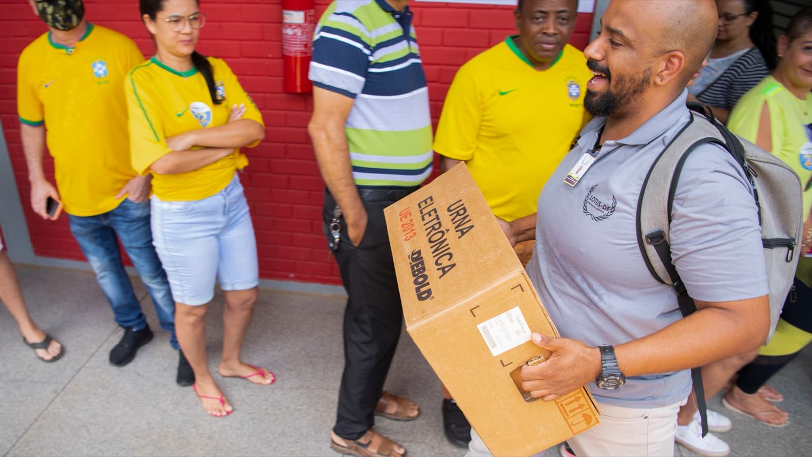 Foto de arquivo mostra funcionário carregando caixa de urna eletrônica em local de votação, com eleitores ao fundo (eleições de 2022)