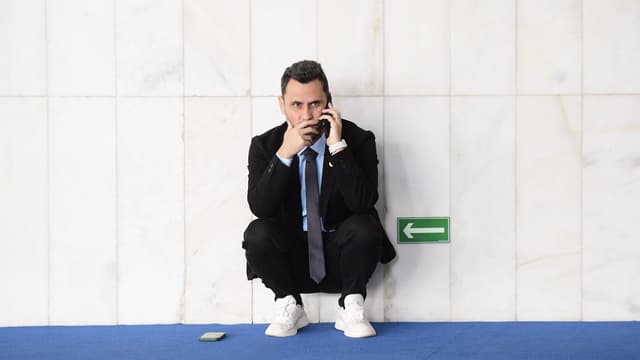 O senador Cleitinho aparece agachado, encostado em uma parede de mármore de um corredor do Senado, falando ao celular; um segundo aparelho está no chão, sobre o carpete azul. Imagem de arquivo.