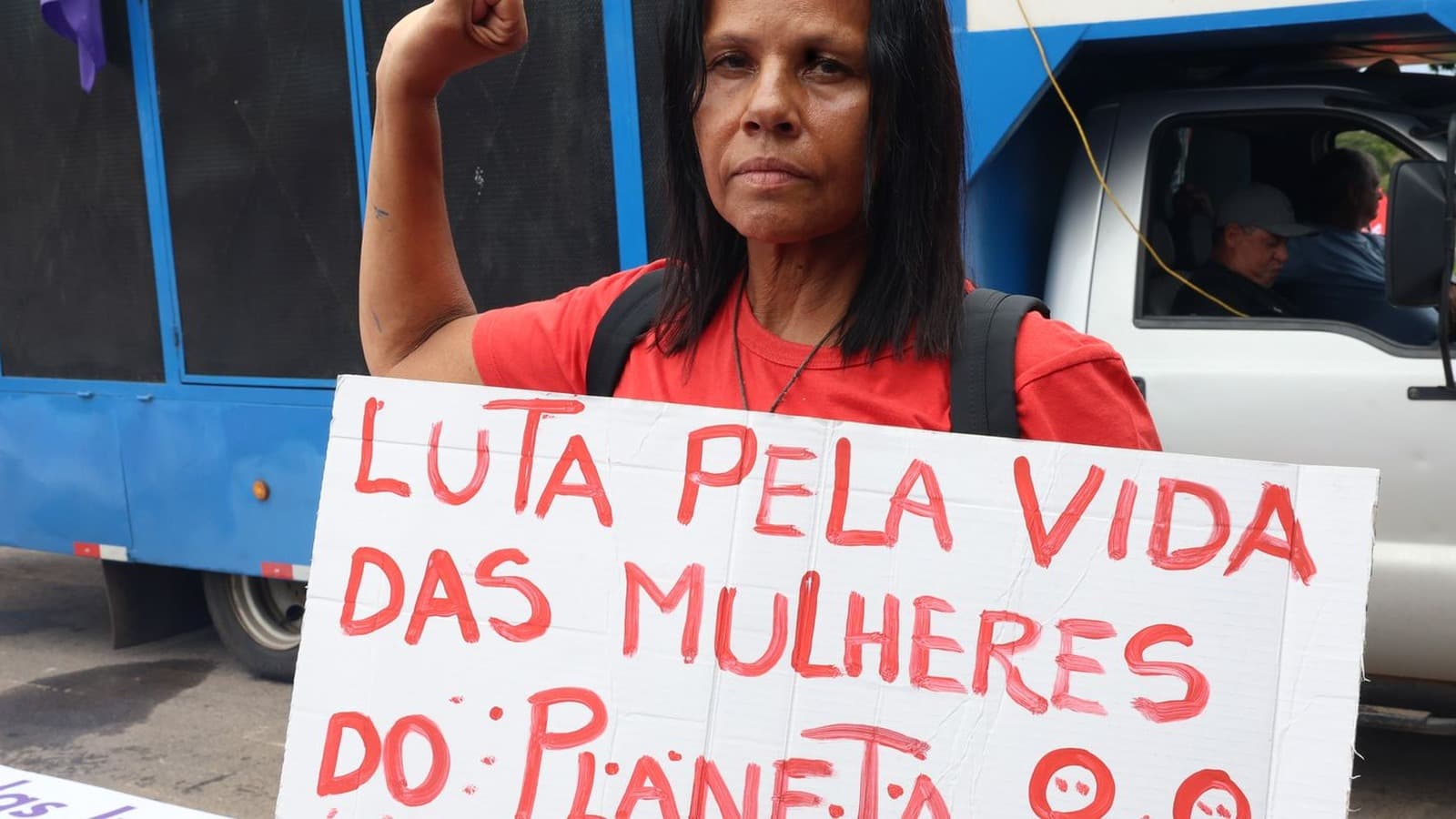 Manifestante de camiseta vermelha com o punho erguido segura cartaz pintado à mão com os dizeres Luta pela vida das mulheres do planeta durante ato de rua; imagem de arquivo