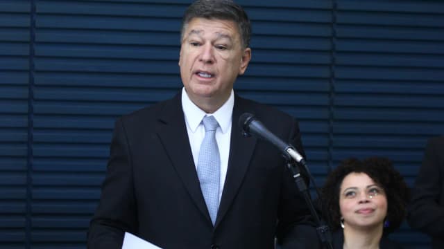 O senador Carlos Viana, de terno escuro, discursa ao microfone em evento de lançamento, com uma mulher sentada ao lado