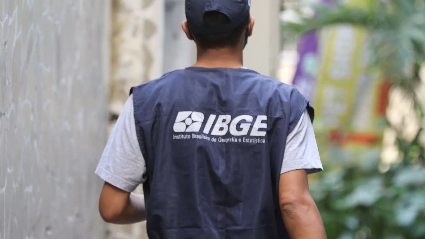 Agente do IBGE visto de costas, vestindo colete com a sigla do instituto, em atividade de campo