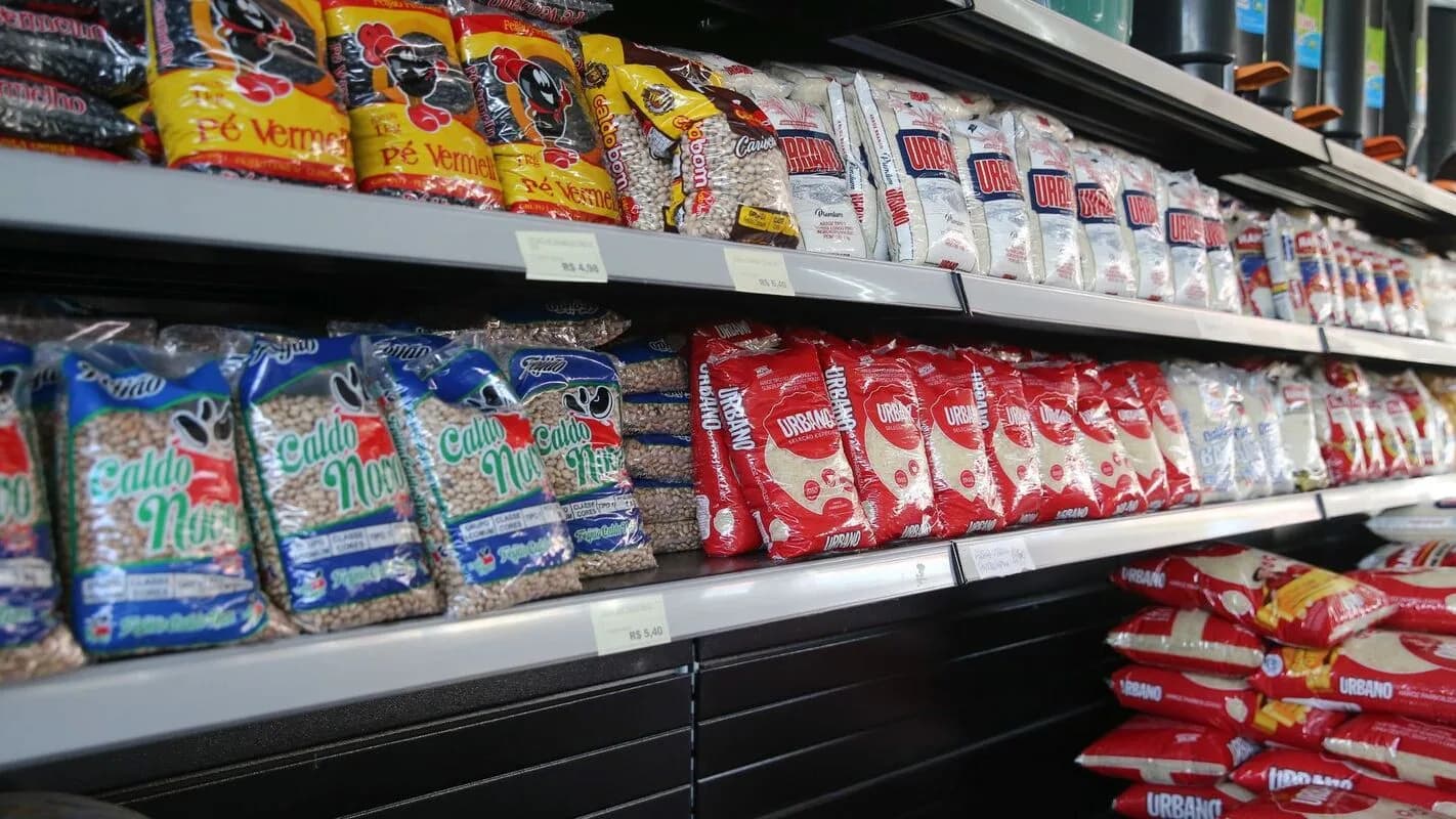 Prateleira de supermercado com pacotes de arroz e feijão, itens da cesta básica