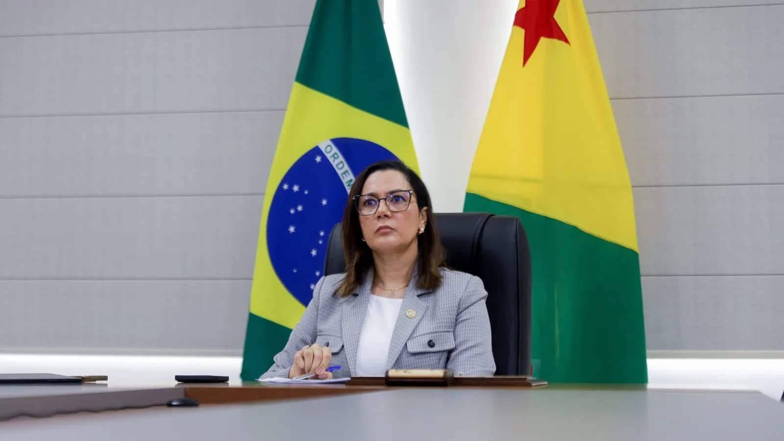 Mailza Assis em ambiente institucional, sentada entre as bandeiras do Brasil e do Acre