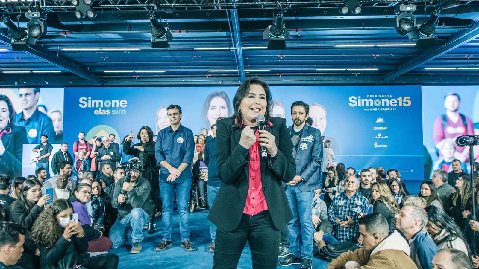 Simone Tebet discursa ao microfone em evento de campanha, com apoiadores ao fundo