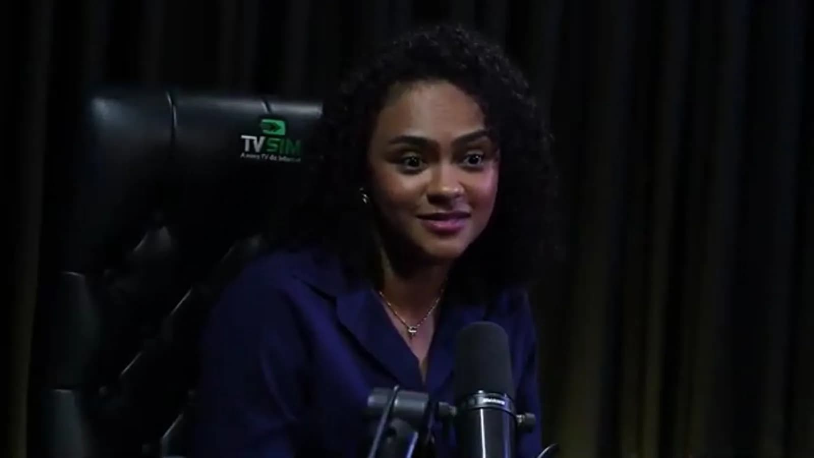 Natália Cordeiro sorri durante entrevista ao Encampos Podcast, sentada em cadeira preta com o logo da TV Sim ao fundo