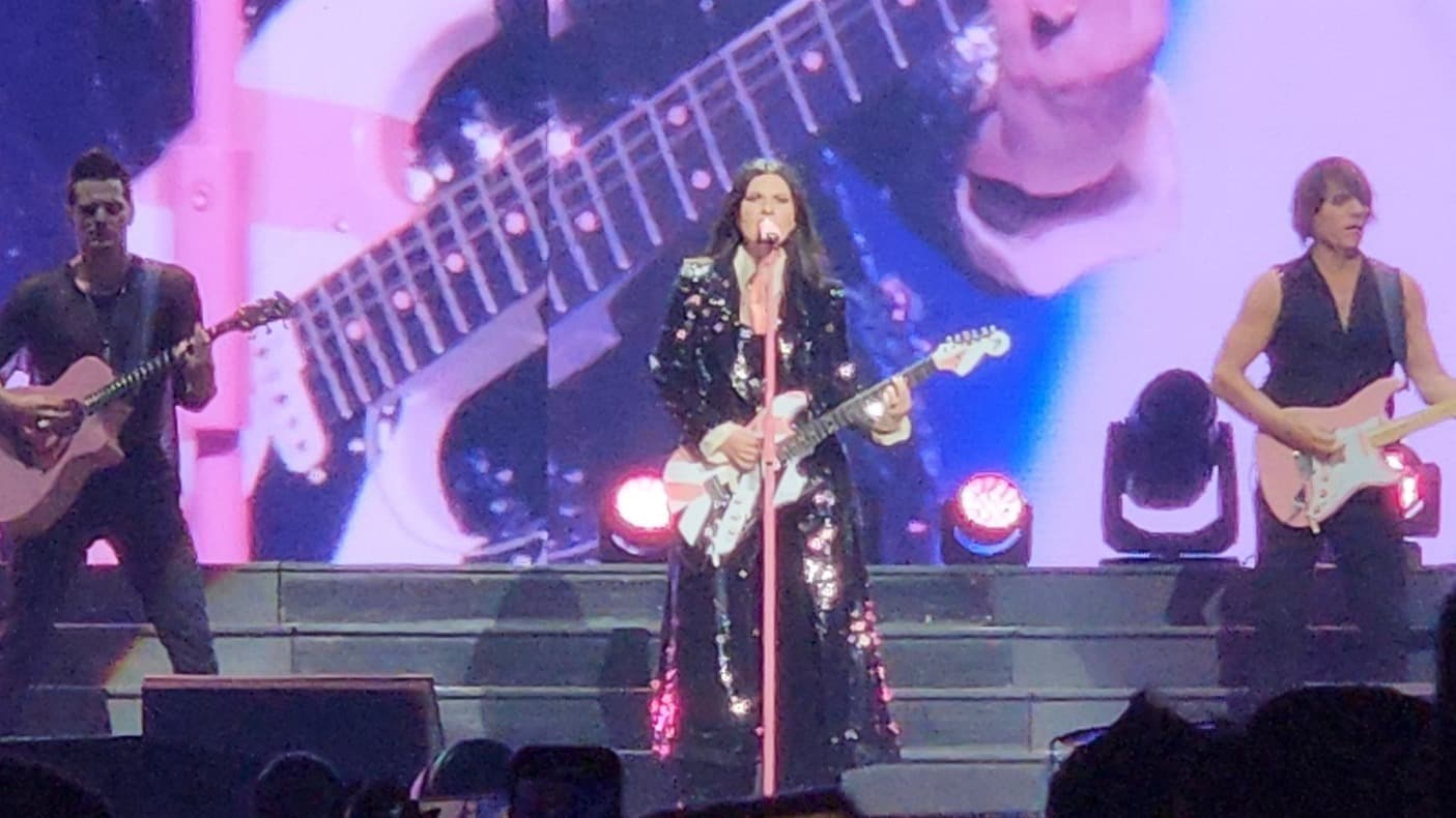 Laura Pausini se apresenta ao microfone durante show, ladeada por guitarristas