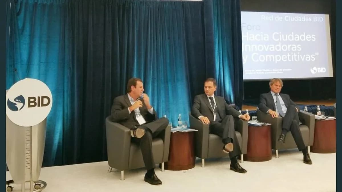 Eduardo Paes fala ao microfone em painel de debate, sentado ao lado de outros dois homens de terno