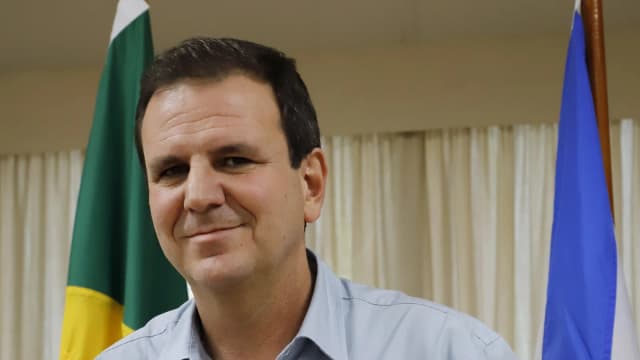 Eduardo Paes sorri em retrato oficial, com bandeiras do Brasil e do Rio de Janeiro ao fundo