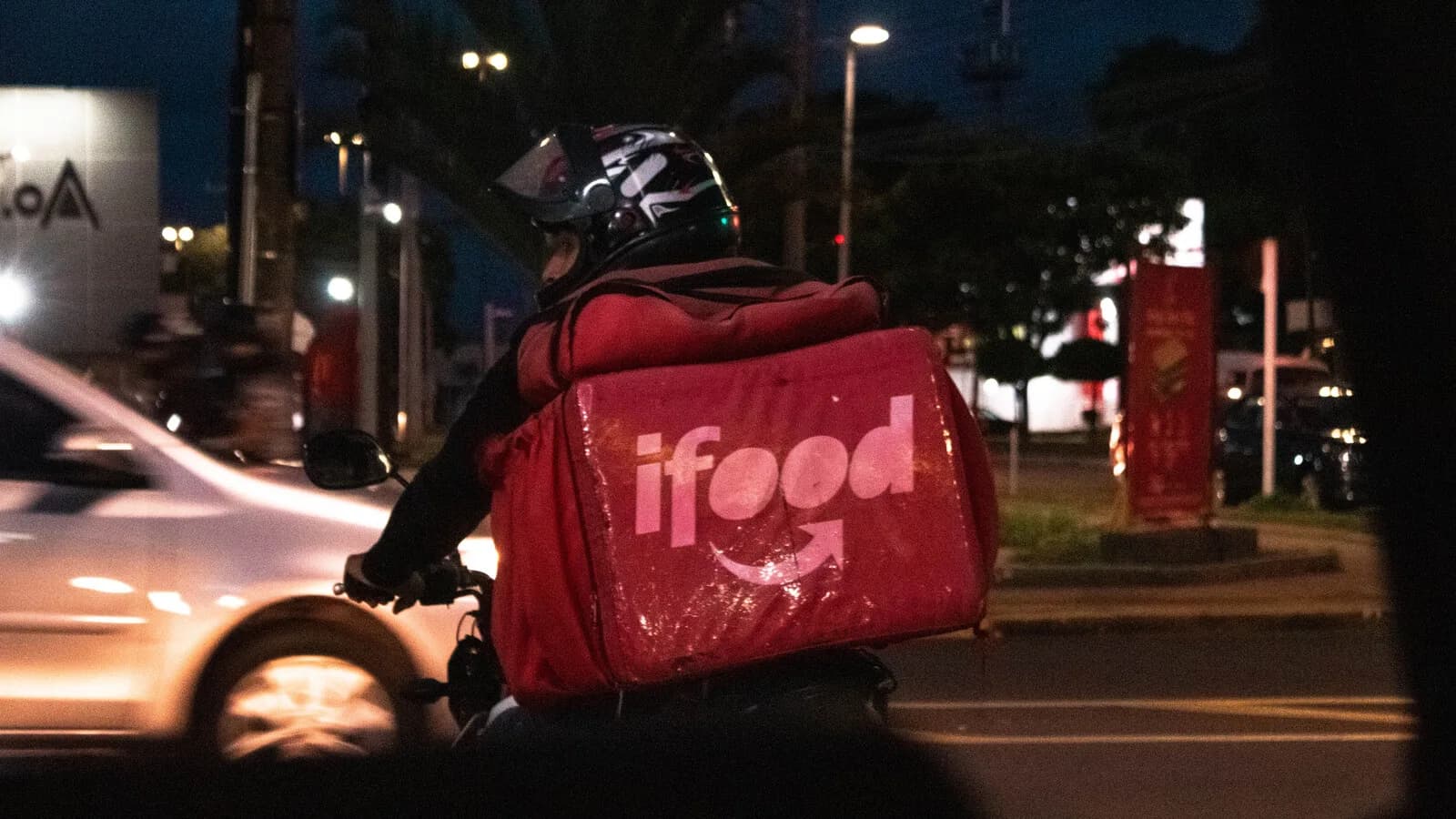 Entregador do iFood em moto à noite, com a bolsa térmica vermelha da empresa, no trânsito de Campo Grande (MS)