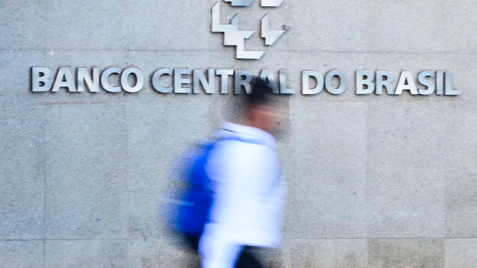 Letreiro metálico com a inscrição Banco Central do Brasil na fachada de granito da sede da autarquia, com um pedestre passando em primeiro plano. Imagem de arquivo