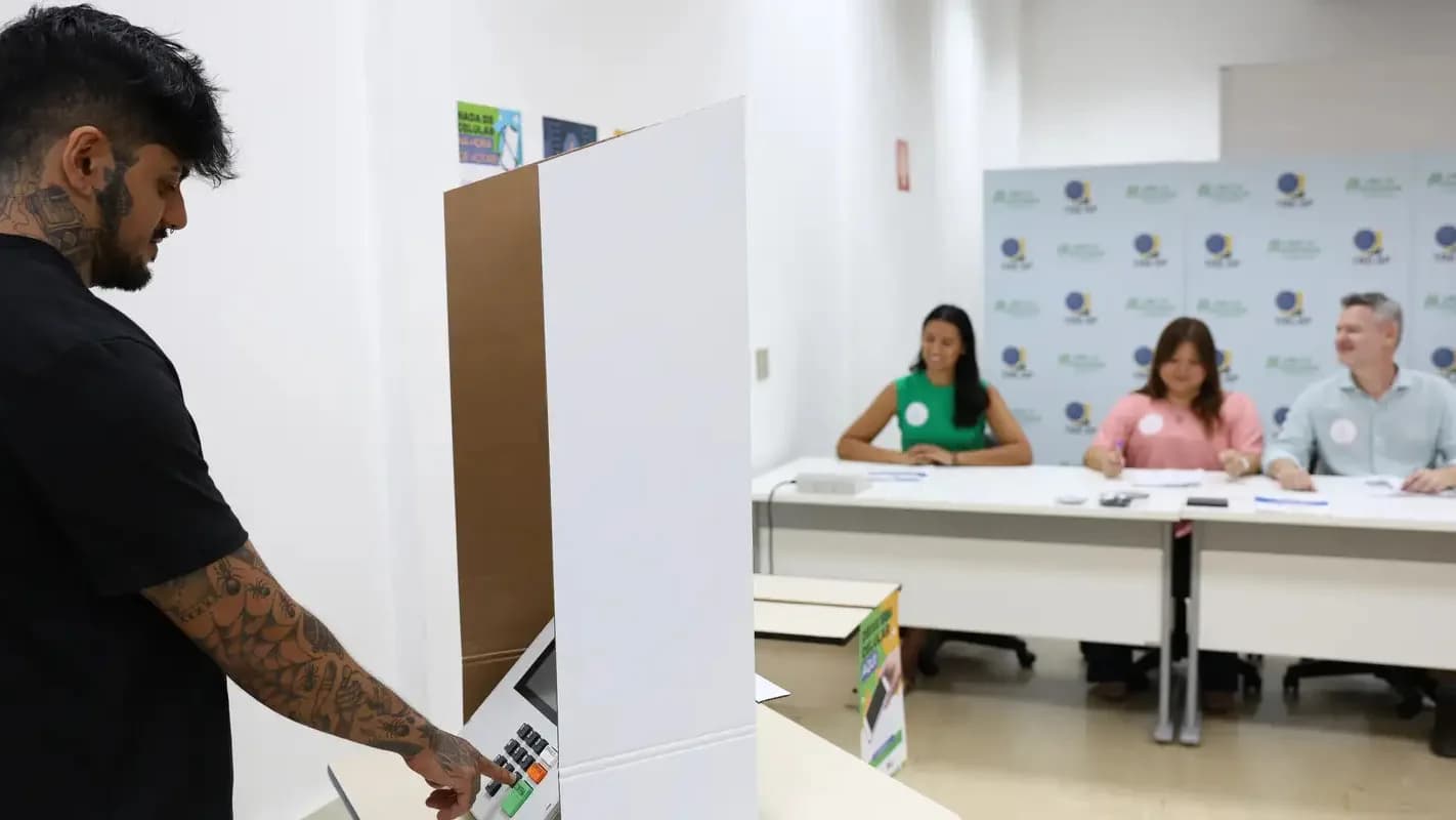 Eleitor vota em urna eletrônica em seção de votação, acompanhado por mesários
