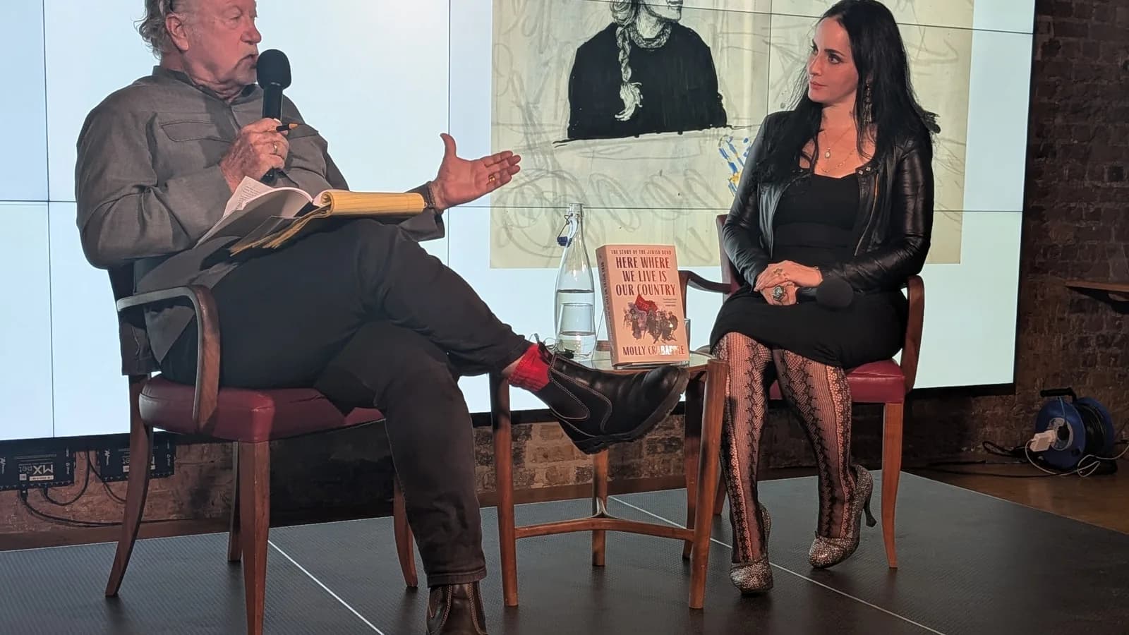 Jon Lee Anderson fala em evento com microfone na mão ao lado de Molly Crabapple